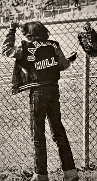 An Oakland Mills student wearing a varsity jacket and holding a pom pom watches a game from a distance through a chain-link fence.